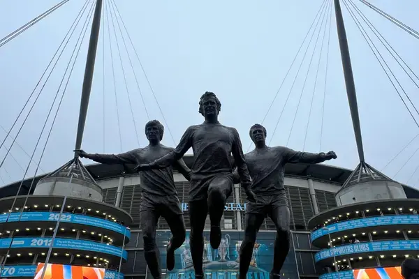 Estátua dos jogadores do Manchester City em frente ao Etihad Stadium, com estrutura metálica e fachada iluminada ao fundo.