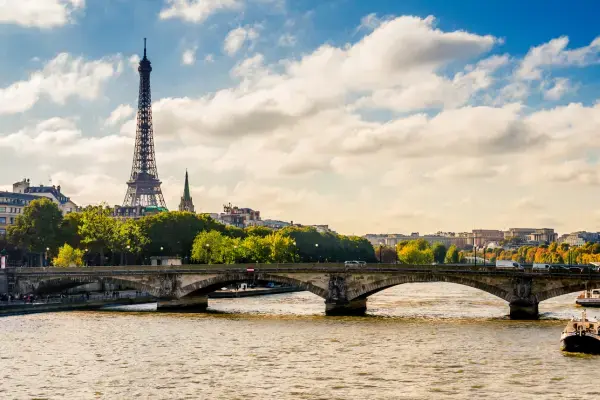 Torre Eiffel ao fundo, com o Rio Sena e uma ponte em primeiro plano, em um dia claro e ensolarado em Paris.