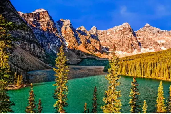 High trees and emerald water in the foreground, mountains covered with white snow and green forest lit by the sun, blue sky with white clouds in the background