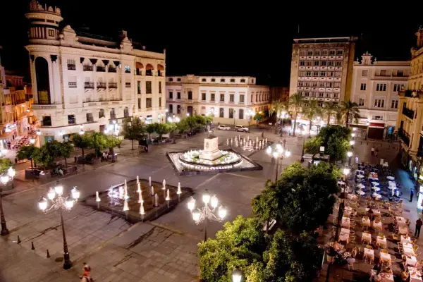 Praça pública em uma cidade iluminada à noite com fonte central, árvores, mesas ao ar livre e edifícios históricos ao redor.