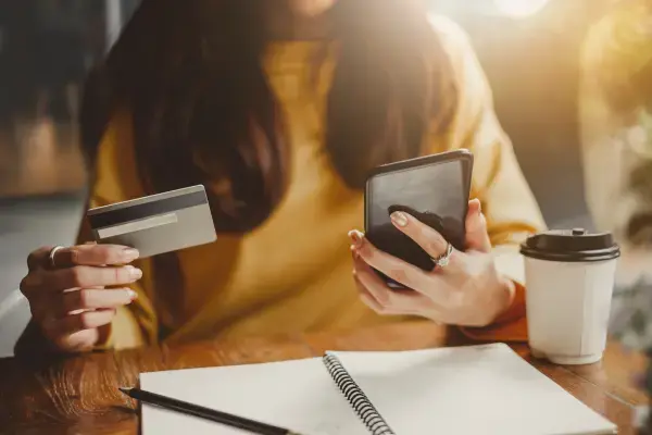 Mulher segurando cartão e celular, com café e caderno na mesa