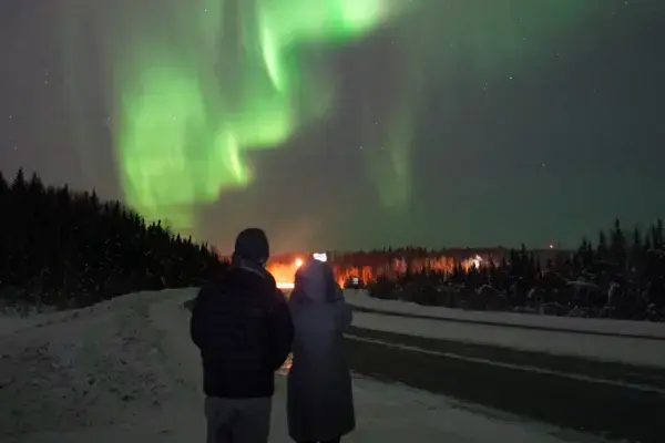 Um casal, visto de costas, observa a Aurora Boreal a partir da beira de uma estrada com neve. O céu exibe luzes esverdeadas intensas acima de uma linha de floresta escura, com luzes distantes no horizonte.