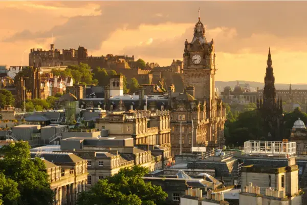 Panorama de Edimburgo com prédios históricos, torres, monumentos e o castelo da cidade iluminado pelo pôr do sol.