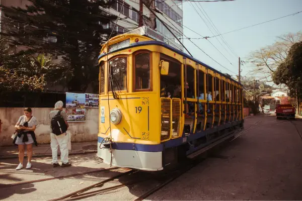 Bondinho amarelo de Santa Teresa passando por uma rua tranquila do Rio, com algumas pessoas ao lado e passageiros dentro.