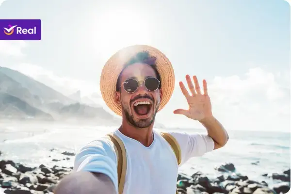 Selfie de um homem muito feliz usando chapéu de palha e óculos escuros. Ele sorri com a boca aberta e acena com a mão para a câmera, tendo ao fundo uma paisagem litorânea ensolarada com pedras escuras e o mar.