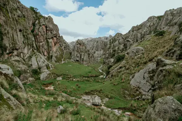Paisagem de um vale rodeado por montanhas rochosas, com gramados verdes e um pequeno rio atravessando a área, sob céu parcialmente nublado.
