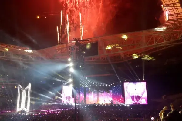 Encerramento do show do BTS no Brasil com fogos de artifício deslumbrantes iluminando o céu noturno. Telões brilhantes e luzes de palco criam uma atmosfera eletrizante e energética.