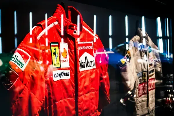 Macacões históricos de pilotos de Fórmula 1 expostos em vitrine, incluindo traje vermelho da Ferrari associado à era Schumacher.