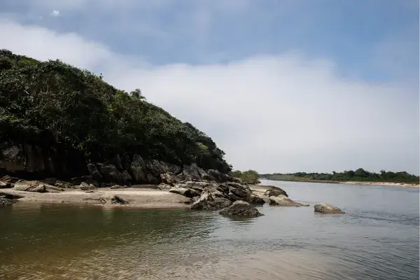 Margem de rio ou canal rodeado por vegetação nativa e pedras, com céu parcialmente nublado e água calma. A paisagem é natural e tranquila, em área preservada do Parque Estadual da Serra do Mar.