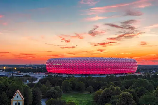 Allianz Arena iluminada em vermelho ao pôr do sol em Munique, com céu alaranjado ao fundo e vegetação ao redor.