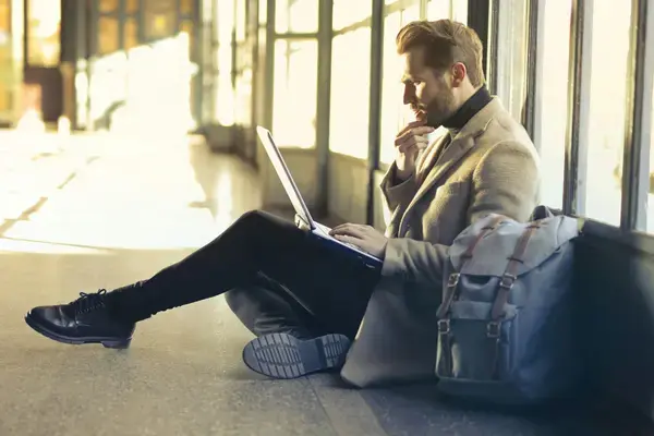 Homem trabalhando no notebook sentado ao lado da mochila de viagem