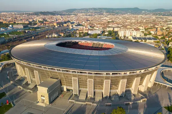Vista aérea da Puskás Aréna iluminada ao entardecer em Budapeste, com as luzes da cidade ao fundo.