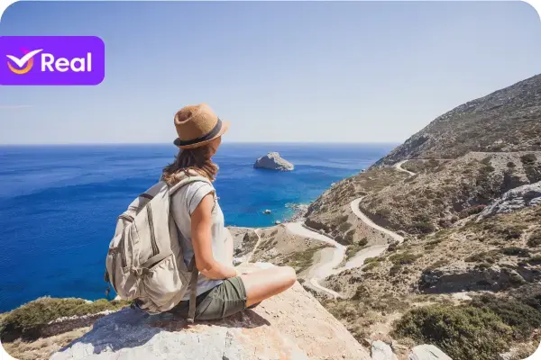 A imagem captura um momento sereno e inspirador de uma viajante solitária em um mirante natural. A cena transmite uma profunda sensação de paz, liberdade e contemplação, emoldurada por uma paisagem costeira espetacular sob um céu azul e ensolarado. A Personagem Principal A figura central é uma mulher, vista de perfil e de costas, sentada de pernas cruzadas sobre uma grande rocha em primeiro plano. Sua postura é relaxada e meditativa, enquanto ela olha para a vasta paisagem à sua frente. Ela está vestida para uma jornada de exploração: Usa uma camiseta clara, shorts em tom de verde militar e um chapéu de feltro marrom estilo fedora, que lhe confere um ar de aventureira moderna. Ao seu lado, uma mochila de viagem grande e de cor clara (bege ou cinza) indica que ela está em meio a uma trilha ou um dia de exploração. A Paisagem Deslumbrante A vista que a viajante contempla é o elemento mais impactante da fotografia: O Mar: Um oceano de um azul profundo e intenso se estende até o horizonte. A luz do sol brilha sobre a superfície calma da água. A Ilha: No meio do mar, uma pequena ilha rochosa se destaca, servindo como um ponto focal na imensidão azul. A Costa e a Estrada: À direita, uma montanha robusta e de vegetação esparsa desce íngrememente em direção ao mar. Cortando a encosta da montanha, uma estrada estreita e sinuosa serpenteia pela paisagem, adicionando uma sensação de jornada e escala à cena. Composição e Atmosfera A fotografia é banhada por uma luz solar intensa e clara, típica de um dia de verão sem nuvens. A paleta de cores é vibrante, dominada pelo azul do mar, os tons terrosos da montanha e das rochas, e as cores neutras da roupa da viajante. A composição, com a mulher posicionada à esquerda e olhando para a vastidão à direita, cria um equilíbrio visual e evoca sentimentos de admiração, introspecção e o prazer de viajar sozinho. Elemento Gráfico No canto superior esquerdo, há um logotipo sobreposto à imagem. É um retângulo roxo com cantos arredondados, contendo um "check" (✓) branco e a palavra "Real", identificando a imagem como material promocional da Real Seguro Viagem.