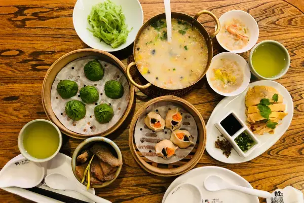 Variedade de pratos de dim sum, incluindo har gow, bolinhos de legumes, chá verde e sopa de canja, servidos em mesa de madeira, estilo chinês.