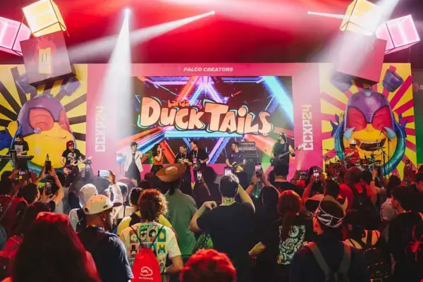 Apresentação ao vivo na CCXP, com uma banda tocando no palco enquanto a plateia assiste e grava. O telão exibe o nome "DuckTails". O ambiente está iluminado com luzes coloridas e há figuras coloridas ao redor do palco.