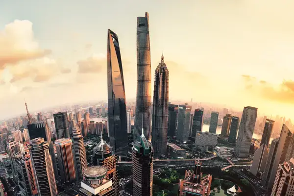 Vista aérea panorâmica de ângulo amplo (fisheye) do distrito financeiro de Pudong, em Xangai, durante o pôr do sol. O horizonte é dominado pelos três arranha-céus mais altos da cidade: a Shanghai Tower, o Shanghai World Financial Center e a Jin Mao Tower, cujas fachadas refletem a luz dourada do sol. Abaixo, a densa malha urbana se estende com inúmeros edifícios comerciais e áreas verdes, sob um céu com nuvens suaves em tons de laranja e amarelo.