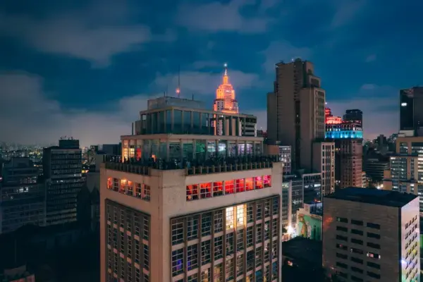 Vista noturna do centro de São Paulo com prédios iluminados. Em destaque, um edifício com rooftop movimentado, luzes coloridas e o Farol Santander ao fundo.