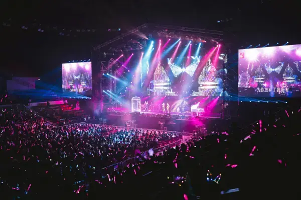 Show em grande arena coberta, com palco iluminado por luzes coloridas em tons de rosa e azul. Telões laterais exibem a performance, enquanto a plateia acompanha de perto com bastões luminosos.