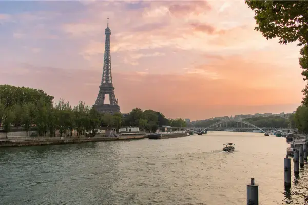 Torre Eiffel ao entardecer vista do Rio Sena, com barco turístico navegando e céu alaranjado.