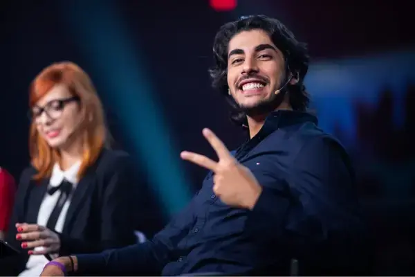 Baiano sorri para a câmera e faz sinal de paz enquanto participa de uma mesa de transmissão de League of Legends, com iluminação de palco e outros comentaristas ao fundo.