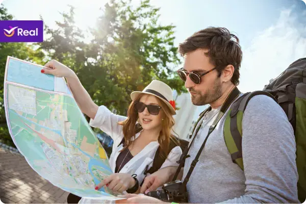 Casal de turistas jovens ao ar livre consultando um mapa de papel grande. O homem usa uma mochila de viagem grande e câmera fotográfica no pescoço, e a mulher usa chapéu e aponta para um local no mapa, ambos iluminados pela luz do sol.