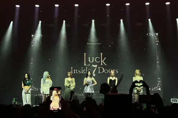 Foto de um show de k-pop em palco iluminado, com seis integrantes do grupo se apresentando diante de um telão que exibe o título “Luck Inside 7 Doors”, enquanto fãs levantam celulares e lightsticks.