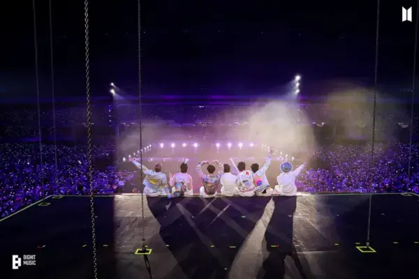 Sete integrantes do BTS em um show ao vivo no estádio, de costas para a câmera, com iluminação brilhante e uma grande multidão na plateia.