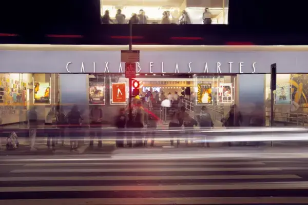 Fachada iluminada do cinema Caixa Belas Artes à noite, em São Paulo. Pessoas aguardam na calçada enquanto carros passam em movimento, deixando rastros de luz.