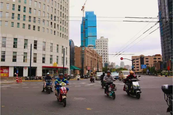O foco central da imagem recai sobre vários condutores de motonetas elétricas ou ciclomotores atravessando um cruzamento amplo. Eles vestem roupas práticas para o dia a dia, incluindo jaquetas e capacetes, refletindo um meio de transporte comum e ágil nas metrópoles chinesas. À esquerda, há um edifício moderno de cor clara com janelas simétricas. No térreo, é visível uma unidade da rede McDonald's, identificada pelo icônico logotipo do "M" amarelo. Ao fundo, o cenário mistura prédios de diferentes épocas e estilos. Um arranha-céu moderno em tons de azul, ainda em construção ou com revestimento reflexivo, destaca-se pela sua altura, ladeado por edifícios comerciais de estilo mais tradicional e cores sóbrias.