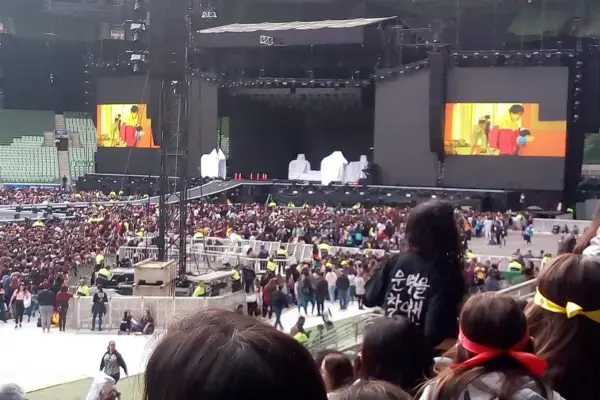 Palco do show do BTS no Allianz Parque do Brasil ao ar livre com dois telões exibindo imagens coloridas. Uma multidão lota o local, criando uma atmosfera vibrante e animada.