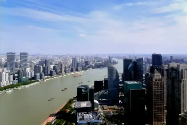 Vista aérea panorâmica do Rio Huangpu serpenteando pela metrópole de Xangai em um dia ensolarado. À direita, destacam-se os modernos arranha-céus de vidro e aço do distrito de Pudong, enquanto à esquerda observa-se a densa malha urbana com edifícios residenciais e comerciais que se estendem até o horizonte sob um céu azul claro com nuvens esparsas. No rio, é possível ver pequenas embarcações de carga navegando suavemente.