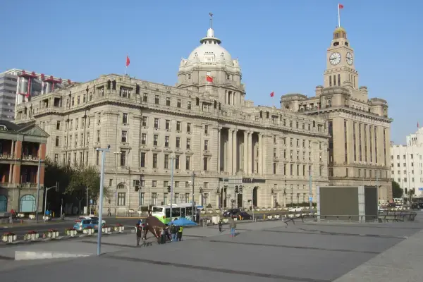 Vista de prédios imponentes de arquitetura neoclássica no Bund, em Xangai. Destacam-se um edifício com uma grande cúpula branca e bandeiras vermelhas, e uma torre de relógio clássica à direita sob um céu azul limpo.