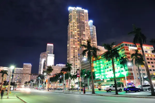 Vista noturna de Miami, com prédios altos iluminados, palmeiras ao longo da avenida e movimento de carros na rua.