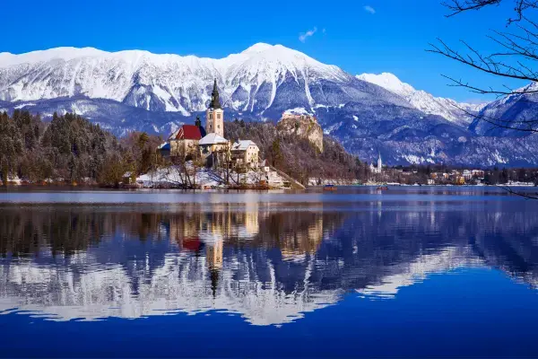 Lago Bled no inverno, com a ilha e sua igreja refletidas na água, e montanhas nevadas ao fundo sob um céu azul.