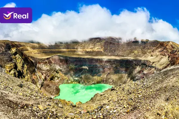 Vista panorâmica da imensa cratera do Vulcão Ilamatepec, em El Salvador, destacando as paredes rochosas e o lago de enxofre cor verde-turquesa no fundo. O céu está azul com algumas nuvens brancas e, no canto superior esquerdo, vê-se a logo roxa da Real Seguro Viagem.