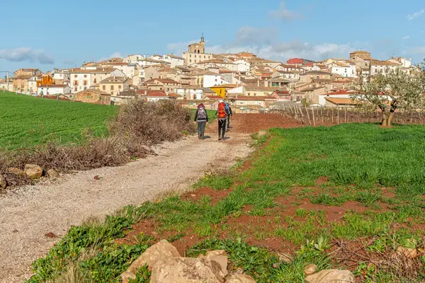Peregrinos caminhando com mochilas em estrada rural rumo a um povoado