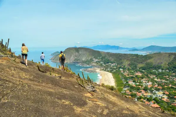 Trilheiros caminhando em pedra com vista panorâmica da praia de Itacoatiara em Niterói, RJ
