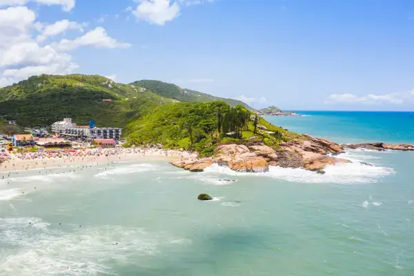 Vista aérea de uma praia em Florianópolis, Santa Catarina, com mar azul, areia clara, formações rochosas e morros cobertos de vegetação ao fundo. A faixa de areia está cheia de banhistas e há construções próximas à orla.