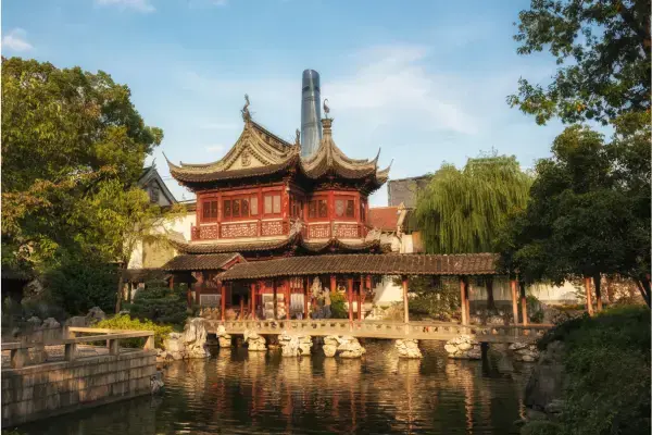 Vista do Yu Garden em Xangai, destacando a arquitetura clássica de um pavilhão com telhados de pontas curvadas e detalhes em madeira vermelha. A estrutura está refletida em um lago calmo cercado por vegetação densa, com uma ponte de pedra coberta em primeiro plano e o contraste da moderna Shanghai Tower ao fundo.