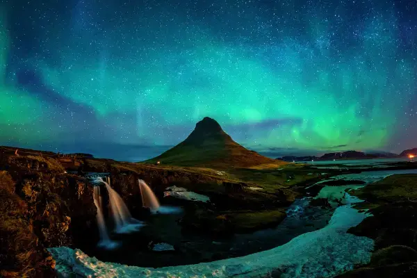 Paisagem noturna icônica mostrando a Aurora Boreal verde sobre a montanha Kirkjufell na Islândia. Em primeiro plano, uma cachoeira flui entre rochas e neve sob um céu estrelado e iluminado pelo fenômeno.