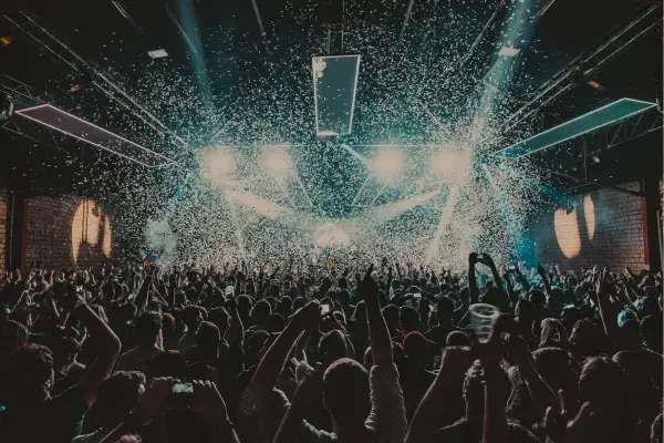 Foto de um show lotado, com o público de braços erguidos enquanto papéis picados caem do teto. No palco, luzes fortes iluminam a multidão, transmitindo a energia da apresentação ao vivo.