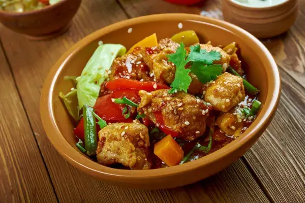 Uma tigela de cerâmica contendo cubos de frango preparados com pimentões vermelhos, cenouras, vagem e sementes de gergelim, decorada com uma folha de coentro fresca.