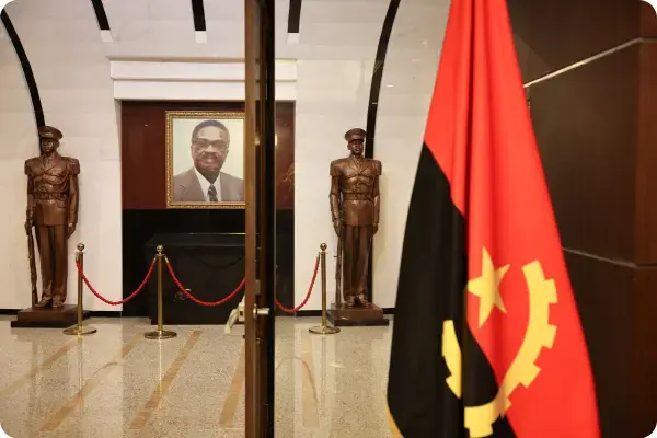 Interior de um memorial oficial com a bandeira de Angola em destaque no primeiro plano à direita. Ao fundo, em uma sala de mármore branco, vê-se um retrato emoldurado de um líder político ladeado por duas estátuas de bronze em tamanho real de soldados em posição de sentido.