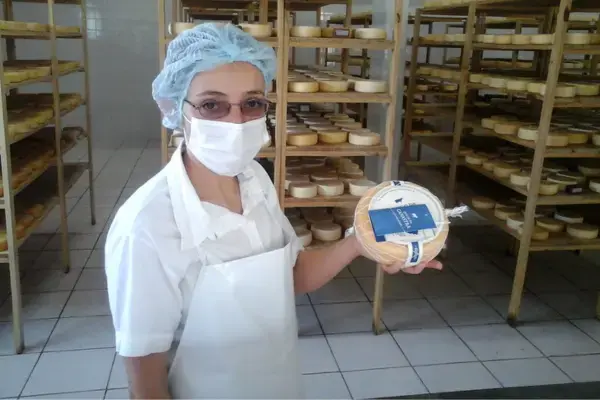 mulher segurando queijo canastra. ela está de máscara e touca branca, ao fundo prateleiras com queijos