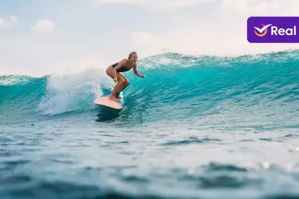 Mulher surfista em ação, dropando uma onda de águas claras e turquesas. Ela usa um maiô escuro e se equilibra na prancha, com o corpo inclinado para a frente. O dia está ensolarado e o mar tem boa formação. No canto superior direito, a logo roxa da Real Seguro Viagem."