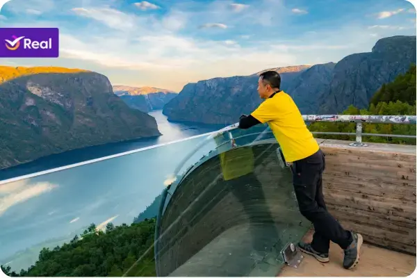 Homem de camisa amarela em pé em um mirante com guarda-corpo de vidro, observando uma vista panorâmica impressionante de um fiorde com águas azuis profundas cercado por montanhas altas e íngremes sob um céu com nuvens.