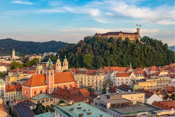 Vista panorâmica de uma cidade com telhados alaranjados, igreja rosa e, ao fundo, um castelo sobre uma colina verde.