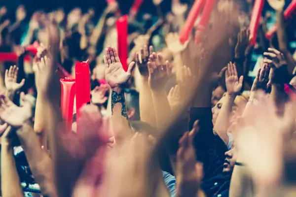 Torcida em estádio com mãos levantadas e bastões infláveis, celebrando durante uma partida de futebol.