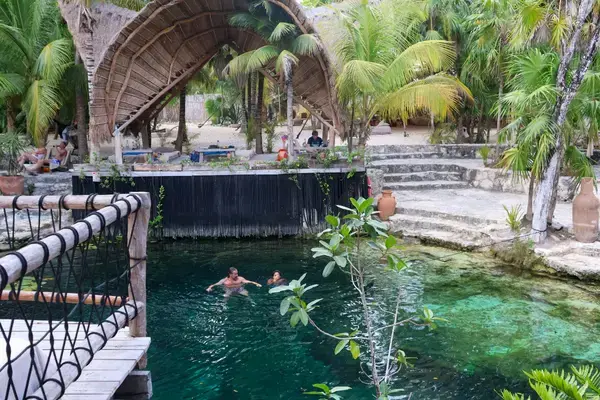 Cenote de águas transparentes com passarela de madeira e vegetação tropical.