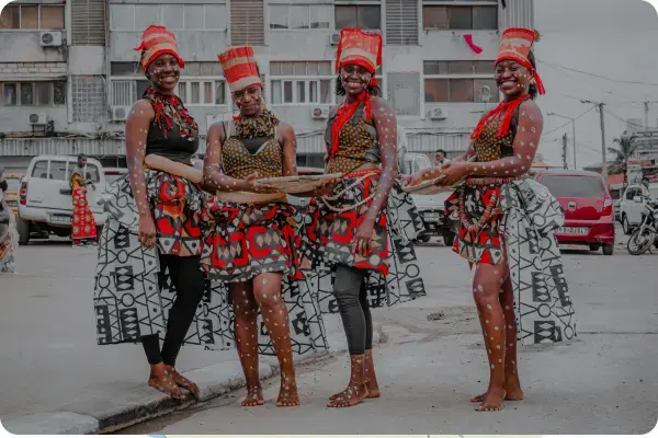 Quatro mulheres angolanas sorridentes posando em uma rua urbana. Elas vestem trajes tradicionais vibrantes com estampas geométricas em preto, vermelho e branco, além de adereços de cabeça cilíndricos. Seus rostos e corpos estão decorados com pequenas marcas brancas rituais.